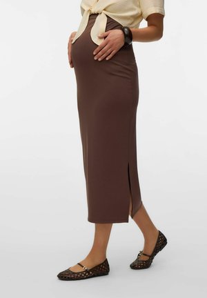 Gravid kvinde iført en beige bundet top, brun tætsiddende nederdel med sidelomme, sort armbånd og sorte flettede flade sko, stående på hvid baggrund.