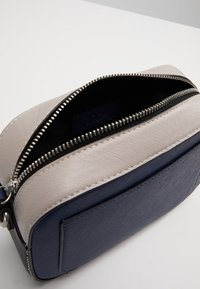 Obdélníková taška s šedým texturovaným víkem, námořnickým modrým dnem a stříbrným zipem. Vnitřní část je vybavena tmavou látkovou podšívkou s etiketou.