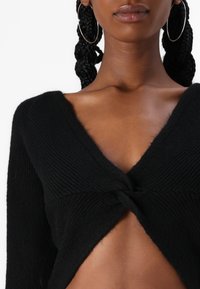 Svart strikket cropped genser med V-hals og vridd frontdetalj, med lange ermer og en myk tekstur.