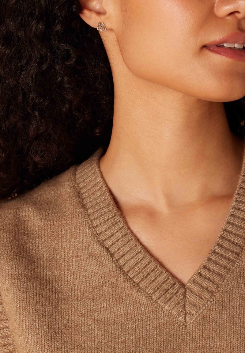 V-hals trui in zachte beige gebreide stof met geribbelde textuur; met daarbij oorbellen met studs in zilver die op elkaar liggende cirkels voorstellen.