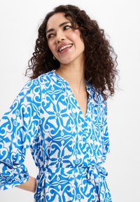 Blauw en wit patroonjurk met een V-hals, lange wijdvallende mouwen en een tailleband. Gemaakt van lichte stof met een gladde textuur.
