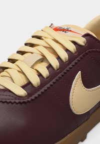 Σκούρο μπορντό δερμάτινο αθλητικό παπούτσι με μπεζ κορδόνια, μπεζ σήμα Nike και κρεμ γλώσσα με πορτοκαλί και μαύρο λογότυπο Nike.