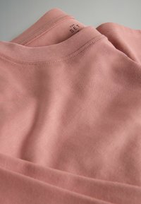 Tricou din bumbac, de culoare roz deschis, cu guler rotund și detalii de cusături vizibile. Materialul pare neted, cu un ușor luciu.