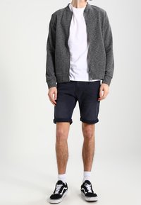Grijze textuur bomberjack over een wit T-shirt, gecombineerd met donkere opgerolde shorts en zwarte sneakers met witte accenten.