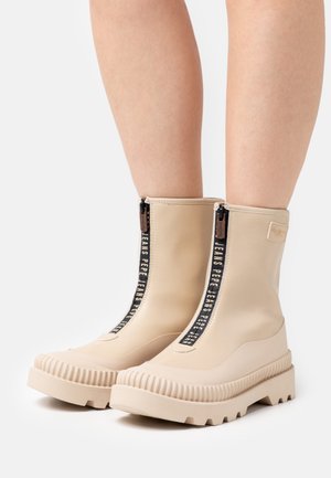 Botas beige hasta la mitad de la pantorrilla con suelas gruesas y acanaladas, cremallera frontal negra con el texto blanco "Pepe Jeans", usadas sobre piernas desnudas contra un fondo blanco.