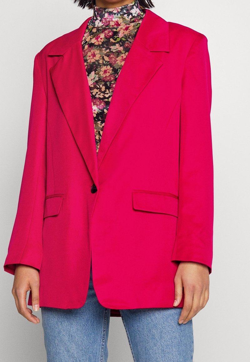 Personne portant un blazer oversize rose vif par-dessus un haut à col montant fleuri et un jean bleu clair, debout devant un fond uni.