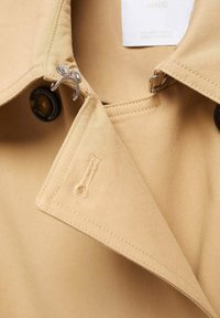 Beżowy kołnierz płaszcza trenchanowego z kontrastującymi czarnymi guzikami, metalowymi elementami i detalami szycia. Posiada gładki, teksturowany materiał.