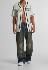 Biała koszula z krótkim rękawem na guziki z czarnym napisem i paskowanymi akcentami, zestawiona z luźnymi ciemnymi dżinsami z dużymi przetarciami.