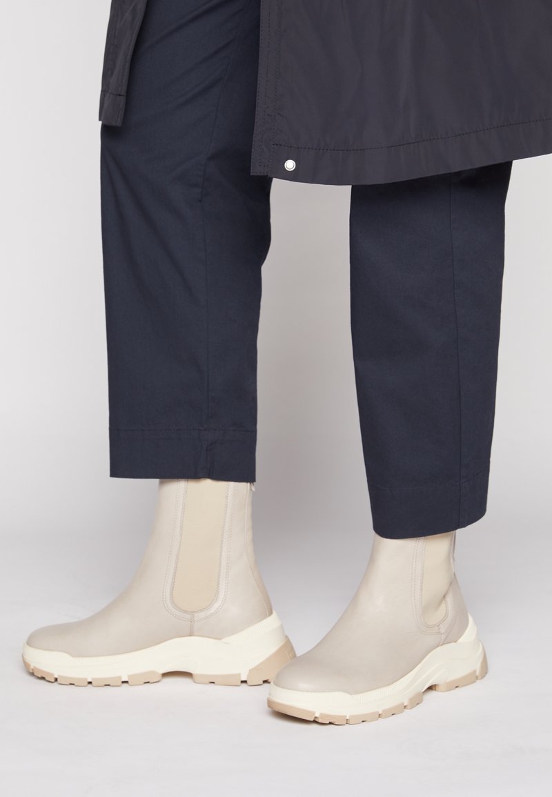 Beige Stiefeletten mit elastischen Seiteneinsätzen, Gummisohlen und strukturierter Oberfläche, kombiniert mit navyblauen, kurz geschnittenen Hosen, die klare Linien und neutrale Farben zur Geltung bringen.