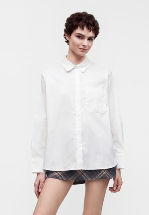 Jeune personne aux cheveux courts et bouclés portant une chemise blanche surdimensionnée boutonnée et une jupe courte à carreaux sur un fond uni.