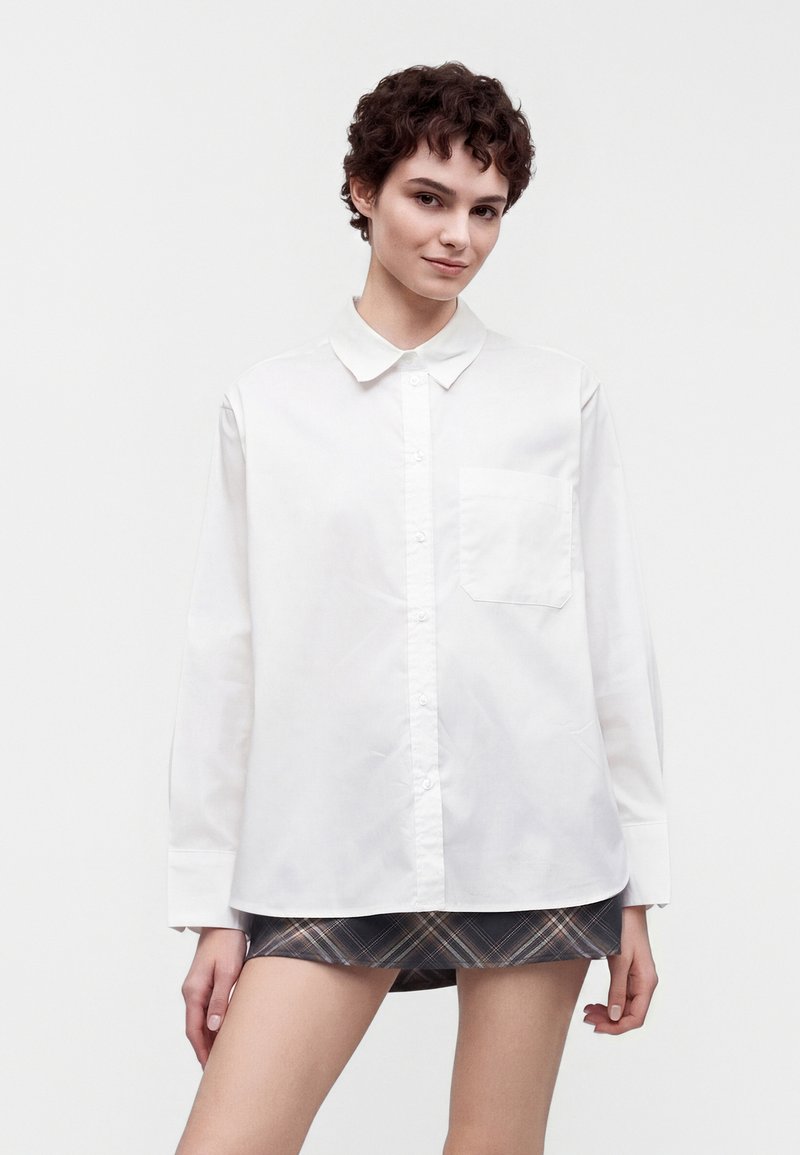 Jeune personne aux cheveux courts et bouclés portant une chemise blanche surdimensionnée boutonnée et une jupe courte à carreaux sur un fond uni.