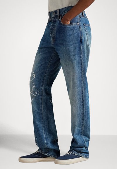 Persona valkā plaša piegriezuma zilas džinsas ar baltām šuvēm un tumši zilas sporta kurpes ar baltu zoli, stāv ar vienu roku kabatā.