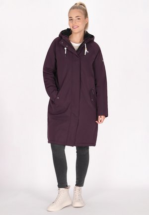 Paarse lange jas met een capuchon, voorzien van ritszakken en een ontspannen pasvorm, gedragen over grijze jeans en lichtbeige sneakers.