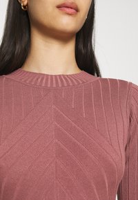 Rosa gerippter Strickoberteil mit einem strukturierten Chevron-Muster und einem runden Halsausschnitt. Nahaufnahme zeigt die Stoffdetails und die Farbe.