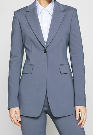 Vrouw draagt een aansluitende grijze blazer met een zwarte knoop en bijpassende grijze broek, daaronder een wit overhemd met kraag.