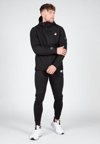 Veste noire zippée avec capuche, dotée d'une petite poche poitrine et d'un logo. Associée à un pantalon noir ajusté et des baskets blanches. Texture lisse.