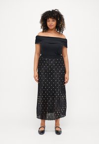 Svart off-shoulder topp kombinert med en svart plissert skjørt prydet med små gullmønstre, fullført med flate svarte sko.