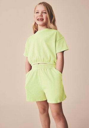 Smilende ung pige med langt blondt hår iført lysegrøn kortærmet top og matchende shorts med hænderne i lommerne mod en beige baggrund.