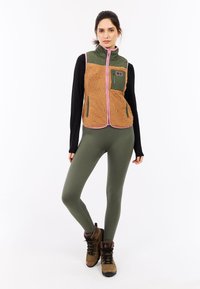 Bruin fleece-voering vest met groene accenten en roze afwerking, gecombineerd met olijfgroene leggings en bruine wandelschoenen.