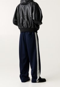 Persona che indossa una giacca di pelle nera, pantaloni da tuta blu navy con strisce bianche sui lati e scarpe nere, in piedi su uno sfondo chiaro e uniforme.
