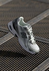 Zapatilla deportiva New Balance en blanco y gris con cordones, situada sobre una superficie de rejilla metálica con iluminación diagonal y patrones de sombras.