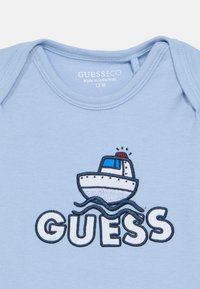 Φωτεινό γαλάζιο βαμβακερό φορμάκι με κεντημένη γραφική απεικόνιση καραβιού πάνω από τη λέξη «GUESS», με σκούρες μπλε γραμμές περιγράμματος και λεπτομέρειες κυμάτων.