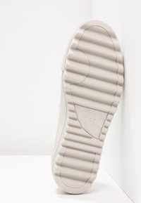 Semelle en caoutchouc blanche de la chaussure Converse All Star avec des rainures texturées et le logo visible au centre de la semelle, reposant contre une surface blanche.