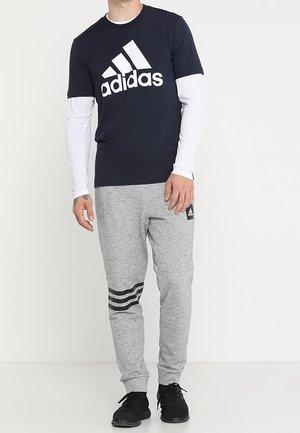 Námořnická modrá tričko Adidas s bílým logem přes dlouhý bílý rukáv; šedé tepláky s černými pruhy a logem na stehně.