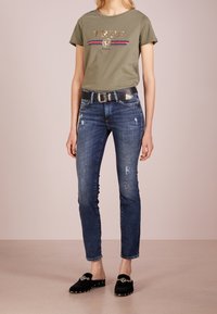 Olivgrön t-shirt med metallisk guldtext, kombinerad med figurnära blå denimjeans med slitna detaljer och ett svart skärp. Svarta slip-on skor.