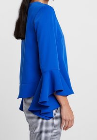 Niebieska bluzka z długim rękawem i rozporkami, gładka w dotyku, z zaokrąglonym dołem, w połączeniu z spodniami w czarno-biały kratę.