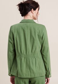 Giacca verde leggera con vita aderente, spalle strutturate e maniche lunghe. Presenta polsini con bottone e una texture sottile.