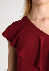 Top burdeos con escote en V, mangas cortas con volantes y textura suave; diseñado con tela de capas para un detalle añadido.