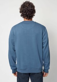 Modrá mikina s dlouhými rukávy, žebrovanými manžetami a kulatým výstřihem. Hladký povrch, minimalistic design, žádné viditelné vzory ani kovové doplňky.