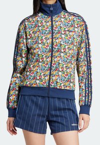 Blommig tryck zip-jacka i flerfärgad tyg med ribbade muddar, parat med shorts i marinblått med ränder. Båda plaggen har logotypdetaljer.