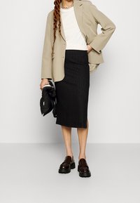 Béžový blazer přes bílý top, černá střihovaná midi sukně a hnědé platformové loafers. Drží černou tašku. Jednoduchý, strukturovaný design.