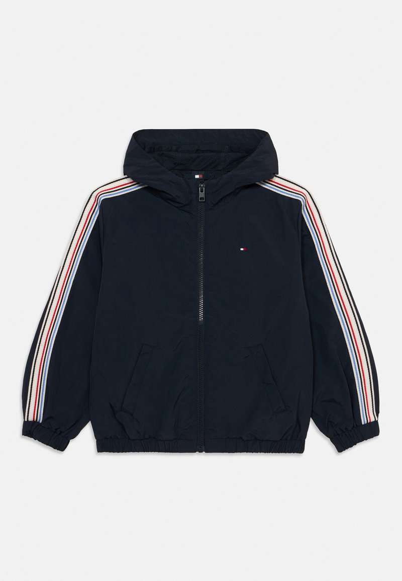 Veste coupe-vent bleu marine avec capuche, présentant des accents rayés blancs, rouges et bleus le long des manches et deux poches avant.