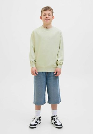 Ljusgrön sweatshirt med texten "Original Studio", tillsammans med blå denimshorts, vita strumpor och svartvita sneakers.