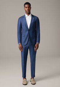 Completo blu a fantasia con un design aderente, abbinato a una camicia bianca. Include polsini arrotolati e mocassini chiari.