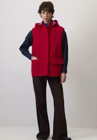 Žena v červenej bezrukávovej kapucňovej veste s veľkými prednými vreckami cez tmavé dlhé rukávy tričko a čierne nohavice, stojí na pozadí bez vzoru.