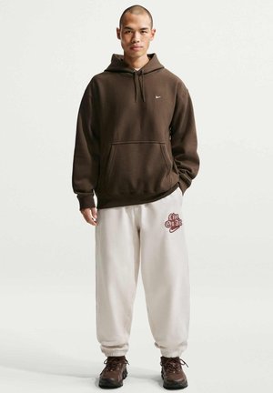 Sweat à capuche marron avec poche avant, cordon de serrage et logo Nike ; pantalon de survêtement blanc avec graphique rouge "Live On Air" ; chaussures noires.