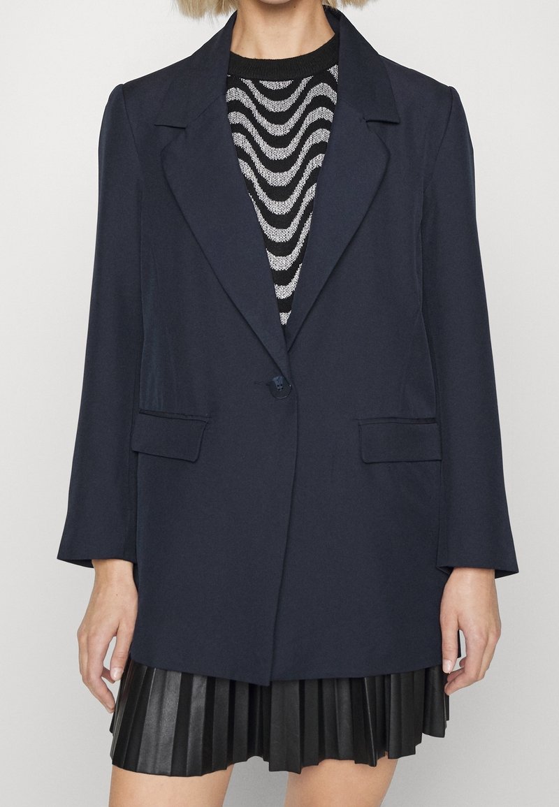 Femme portant un blazer bleu marine par-dessus un pull à motif ondulé noir et blanc et une jupe plissée noire sur un fond uni.