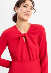 Vrouw glimlachend, gekleed in een rode jurk met lange mouwen en een grote strikdetail bij de halslijn, en zilveren oorringen in de vorm van hoepels.