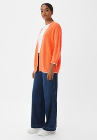 Helles oranges, leichtes Strickcardigan mit lockerem Schnitt und offenem Vorderteil, kombiniert mit einem weißen Top und breiten, dunkelblauen Jeans.