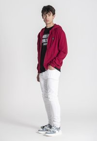 Crvena zip-up majica preko crne majice s grafikom, uparena s bijelim trapericama i tenisicama s plavim detaljima, prikazuje ležeran stil ulične mode.