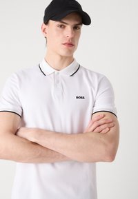 Biely polokošeľa s čiernym lemovaním, na ľavej strane hrude je umiestnené logo. Model má prekrížené ruky a nosí čiernu čiapku. Mäkká textúra.