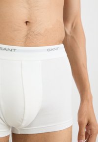 Boxeri din bumbac alb cu un elastic gri pe care este inscripționat logo-ul GANT. Textură netedă și design ajustat, cu detalii vizibile ale cusăturilor.