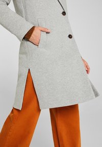 Persona vestida con un abrigo abotonado gris claro y la mano en el bolsillo, combinado con pantalones de pierna ancha de color naranja quemado.
