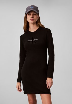 Γυναίκα που φοράει ένα μαύρο φόρεμα Calvin Klein με μακριά μανίκια και ένα σκούρο γκρι καπέλο baseball Calvin Klein, στέκεται μπροστά σε απλό γκρι φόντο.