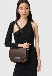 Brązowa skórzana torba crossbody o gładkim wykończeniu, prostokątnym kształcie i metalowym akcentem z przodu. Regulowany pasek, minimalistyczny design.