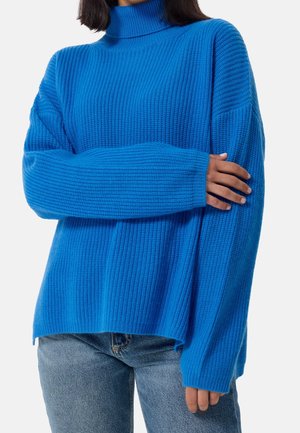 Personne portant un pull à col roulé côtelé bleu vif à manches longues et un jean bleu clair, les bras croisés au niveau de la taille.